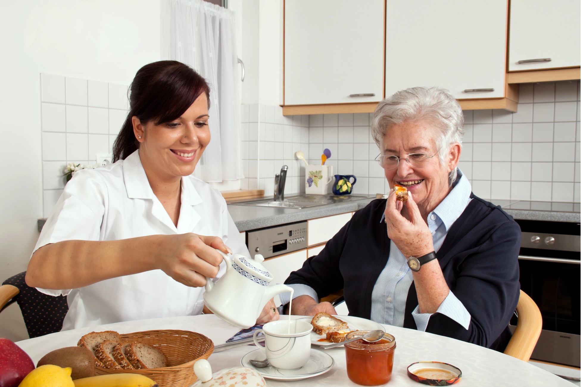 Caregiver pouring tea for elderly woman at breakfast table.